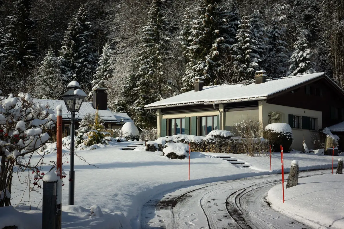 A dark house standing in deep snow, with a person in a red jacket walking away from the camera.